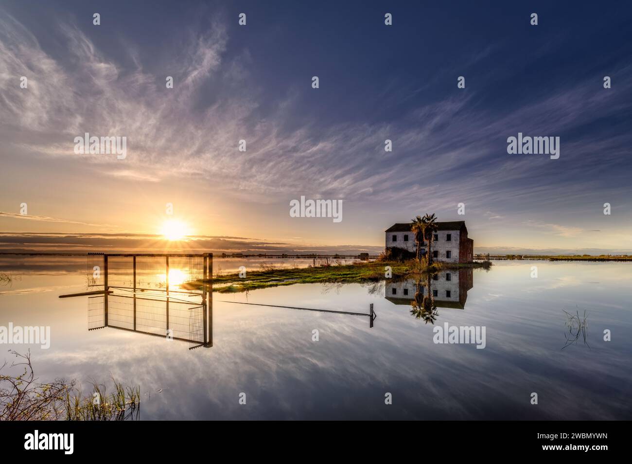 Sunset on the rice fields and flooded road with a rural house with the sun hiding on the horizon and all reflected in the calm water, Sueca, Valencia, Stock Photo