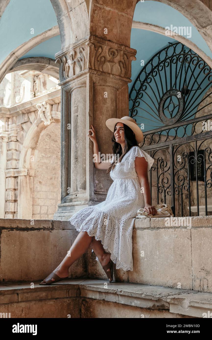A stylish woman sporting a fashionable hat is seen perched gracefully on a ledge of a building, exuding an air of confidence and sophistication Stock Photo