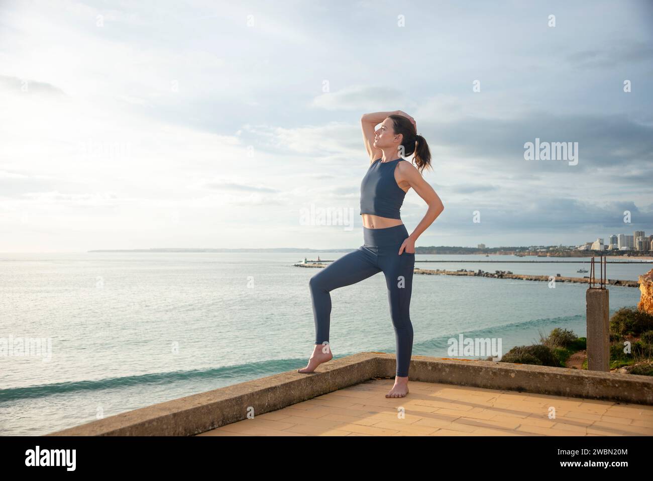 Fit, sporty woman standing by the sea with hand on head, breathing exercise while enjoying the view Stock Photo