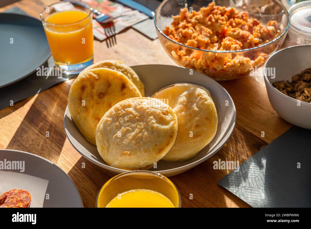 Arepas in the center of the breakfast table next to scrambled eggs, meat and orange juice in direct sunlight. Stock Photo