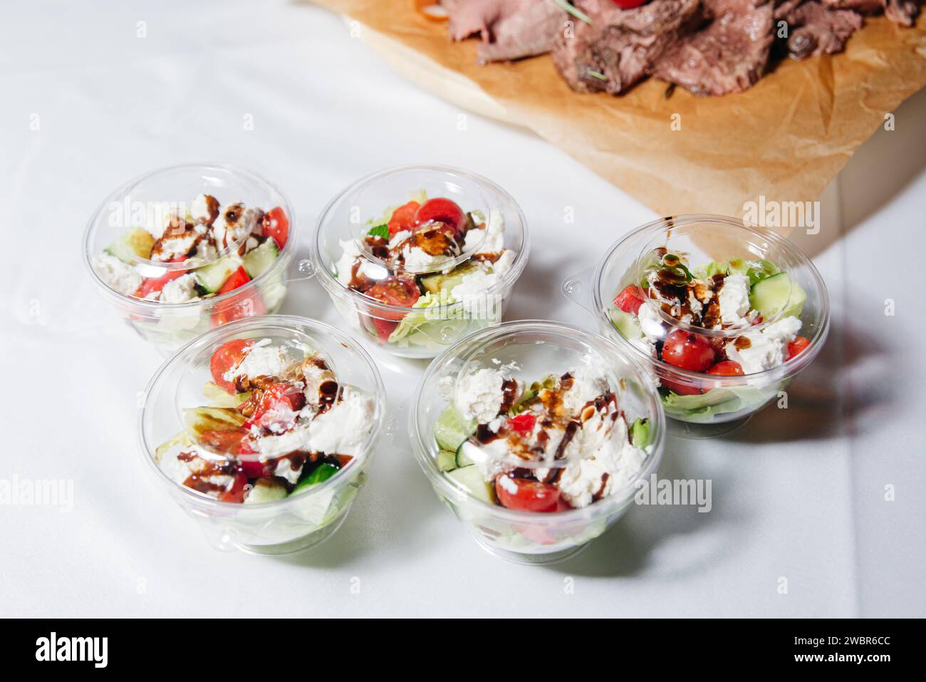 Individual serving bowls of Greek salad with feta cheese, olives, and fresh vegetables, drizzled with balsamic dressing on a white tablecloth. Stock Photo