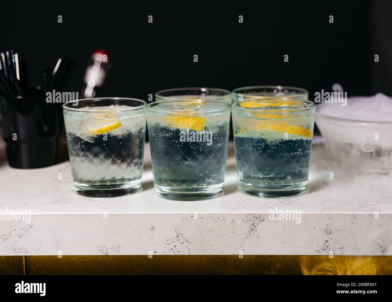 Four mysterious smoky cocktails served on a marble bar counter, each garnished with a slice of lemon, in a dark atmospheric setting. Stock Photo