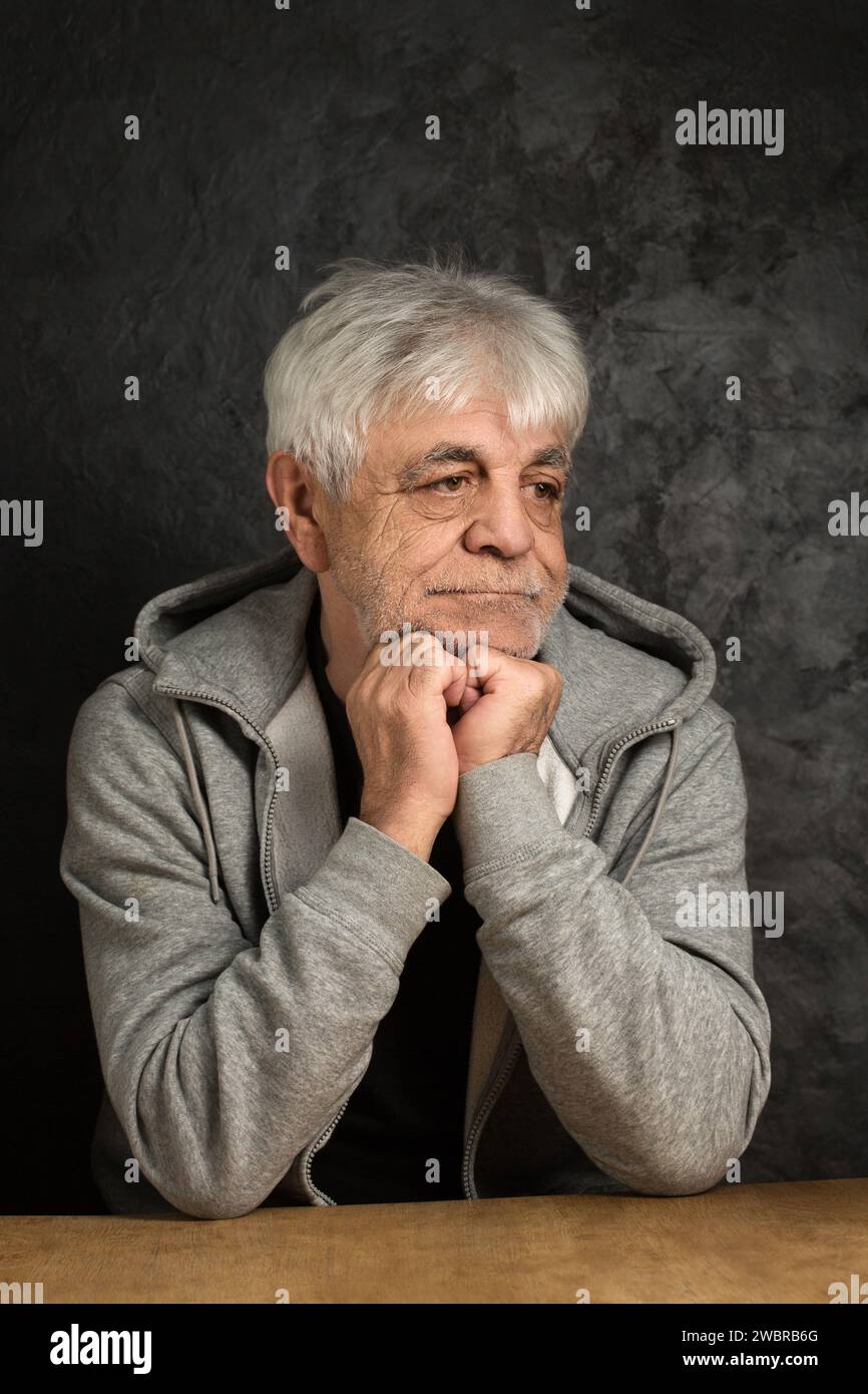 An old man in a hoodie with gray hair is thoughtfully at the table Stock Photo