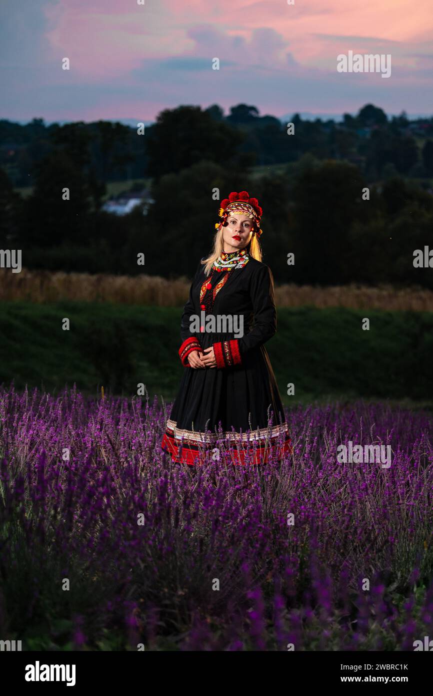 A girl in a chelsea headdress and a black dress decorated with red embroidery stands between lavender bushes. Stock Photo