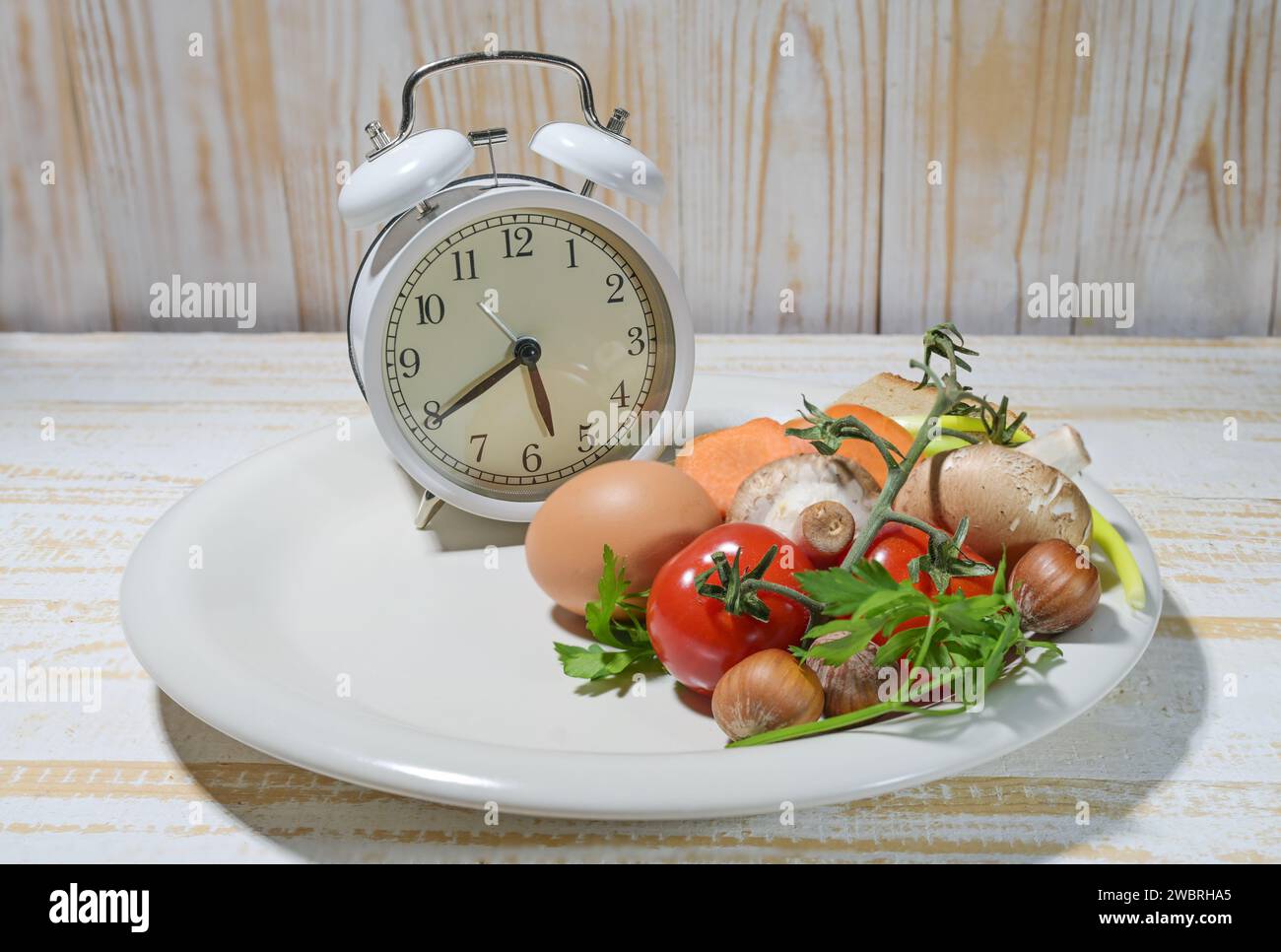 Interval fasting as a healthy diet concept for losing weight, white plate filled to one third with food and an alarm clock on a light rustic wooden ba Stock Photo