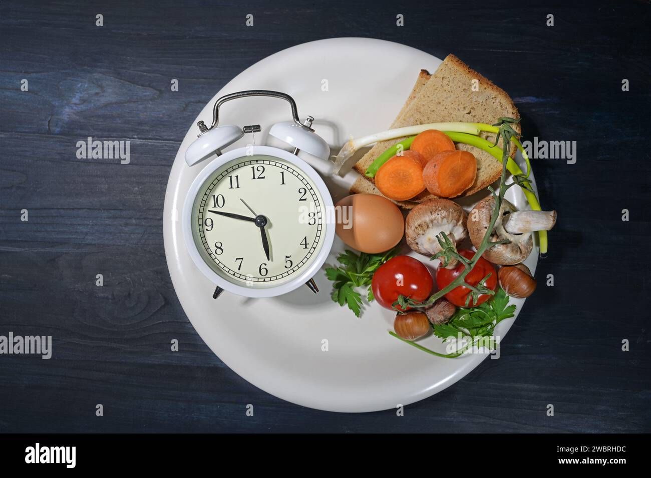 Intermittent fasting, white plate with an alarm clock and partly filled with food on dark blue wood, diet method of eating within a time limit, health Stock Photo