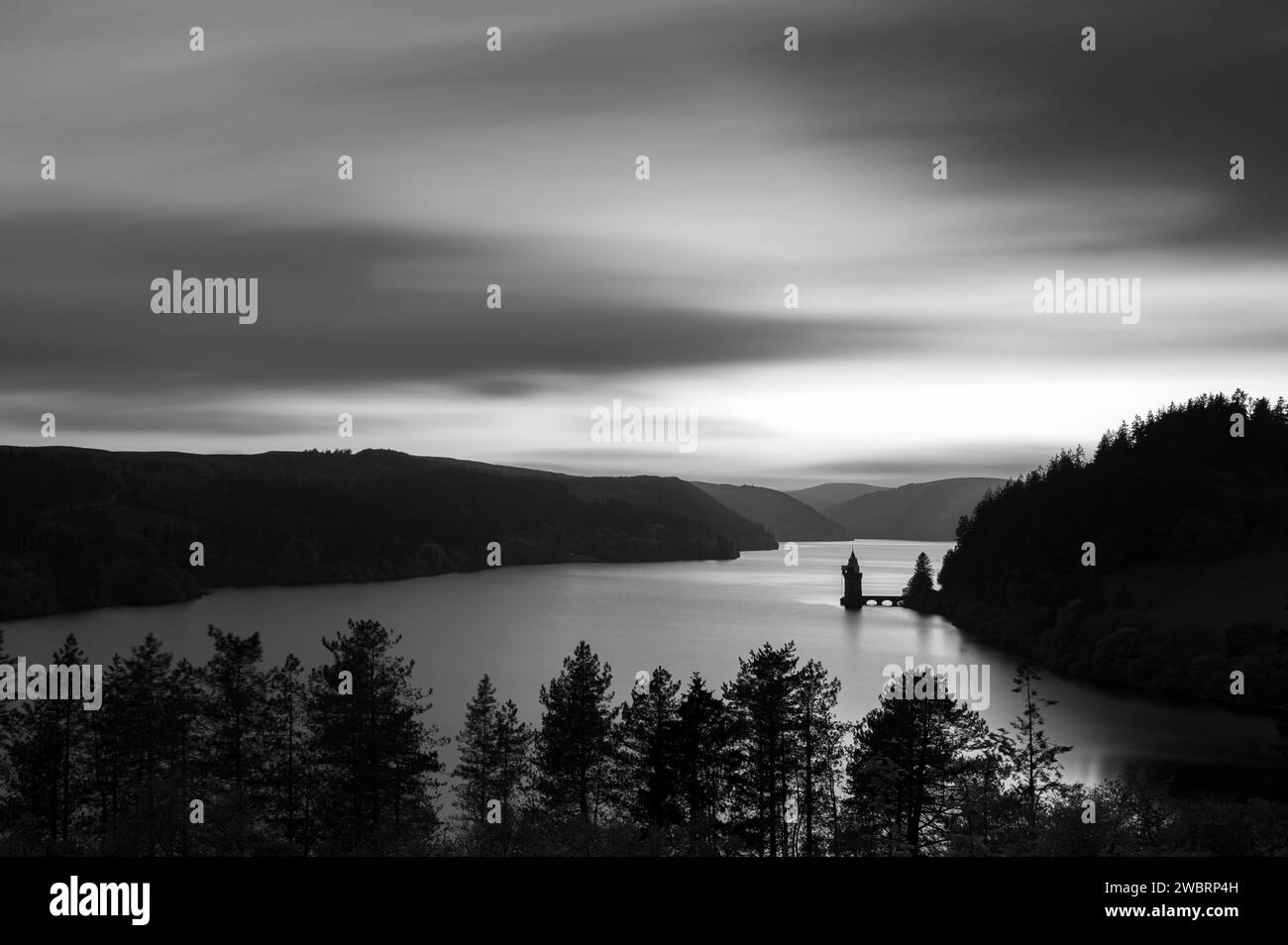 Long exposure, black and white, of Lake Vyrnwy, in Mid Wales. There is a pumping station jutting out into the lake, whcih is surrounded by lush, green Stock Photo