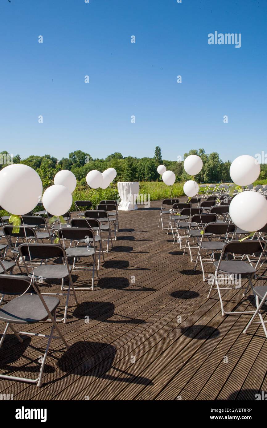 This image captures the tranquil setup of an outdoor ceremony by a lake, before the guests arrive. The scene is composed of rows of metal chairs carefully arranged on a wooden deck, each adorned with a white balloon, adding a touch of festive elegance. The balloons catch the light and sway gently in the breeze, creating a playful yet serene atmosphere. At the focal point of the arrangement stands a solitary podium, draped in white cloth, awaiting the arrival of the speaker or couple. The clear blue sky and lush greenery in the background complete the picturesque setting for what promises to be Stock Photo