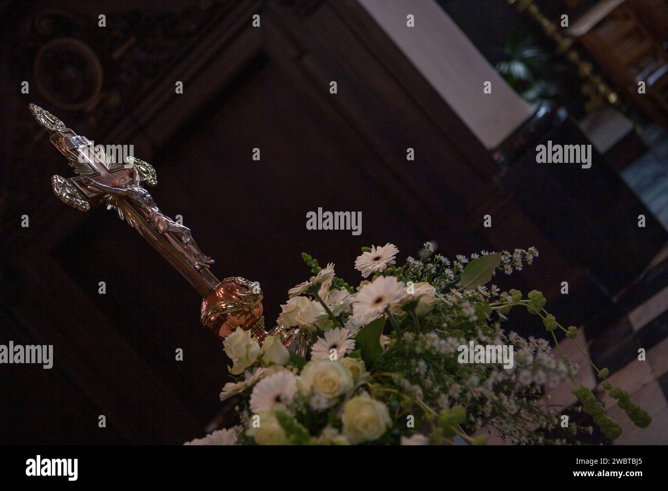 The image captures a powerful religious symbol, a metallic cross, rising above a spray of white flowers, possibly during a solemn church service or ceremony. The cross, with its intricate design, gleams with a reflective sheen, suggesting it is made of brass or a similar material. It stands out against the dark wooden backdrop, typical of a church's rich interior. The flowers, likely part of an altar arrangement, bring a soft, peaceful contrast to the metallic strength of the cross. Sacred Symbolism: The Cross Amidst Floral Peace. High quality photo Stock Photo