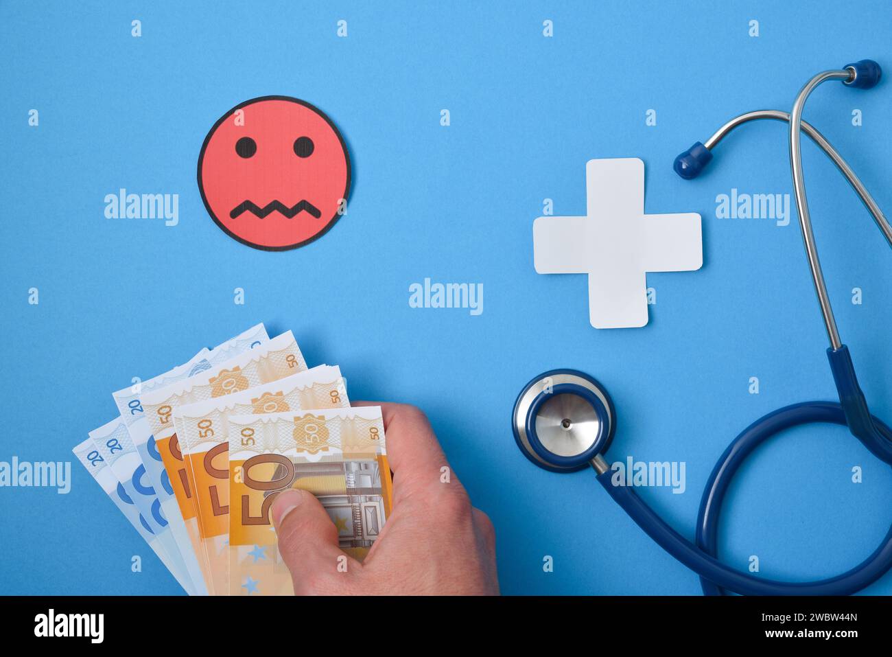 General disagreement with costs in medicine with hand full of money and emoticon and paper cross and stethoscope on blue background. Top view Stock Photo
