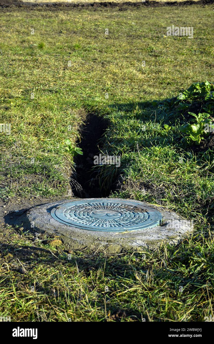 Hatch of well is closed with round metal cover. Sewer well with manhole. Trench is dug to well on green lawn. Grass grows around hatch. Close-up. Sele Stock Photo