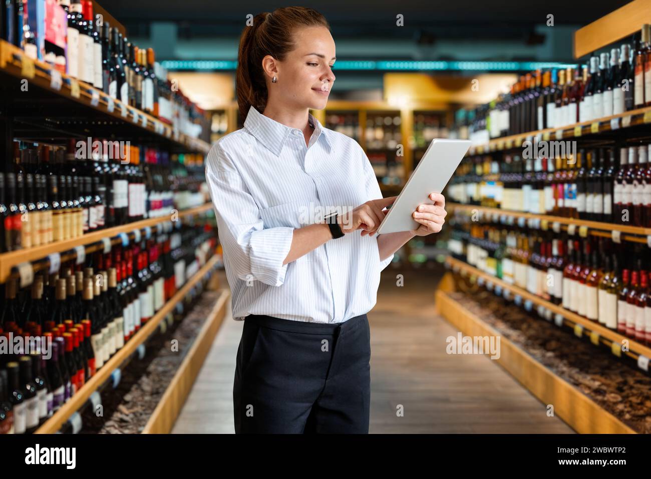 Businesswoman retail manager working on her digital tablet while standing in the wine shop sales area. Stock Photo