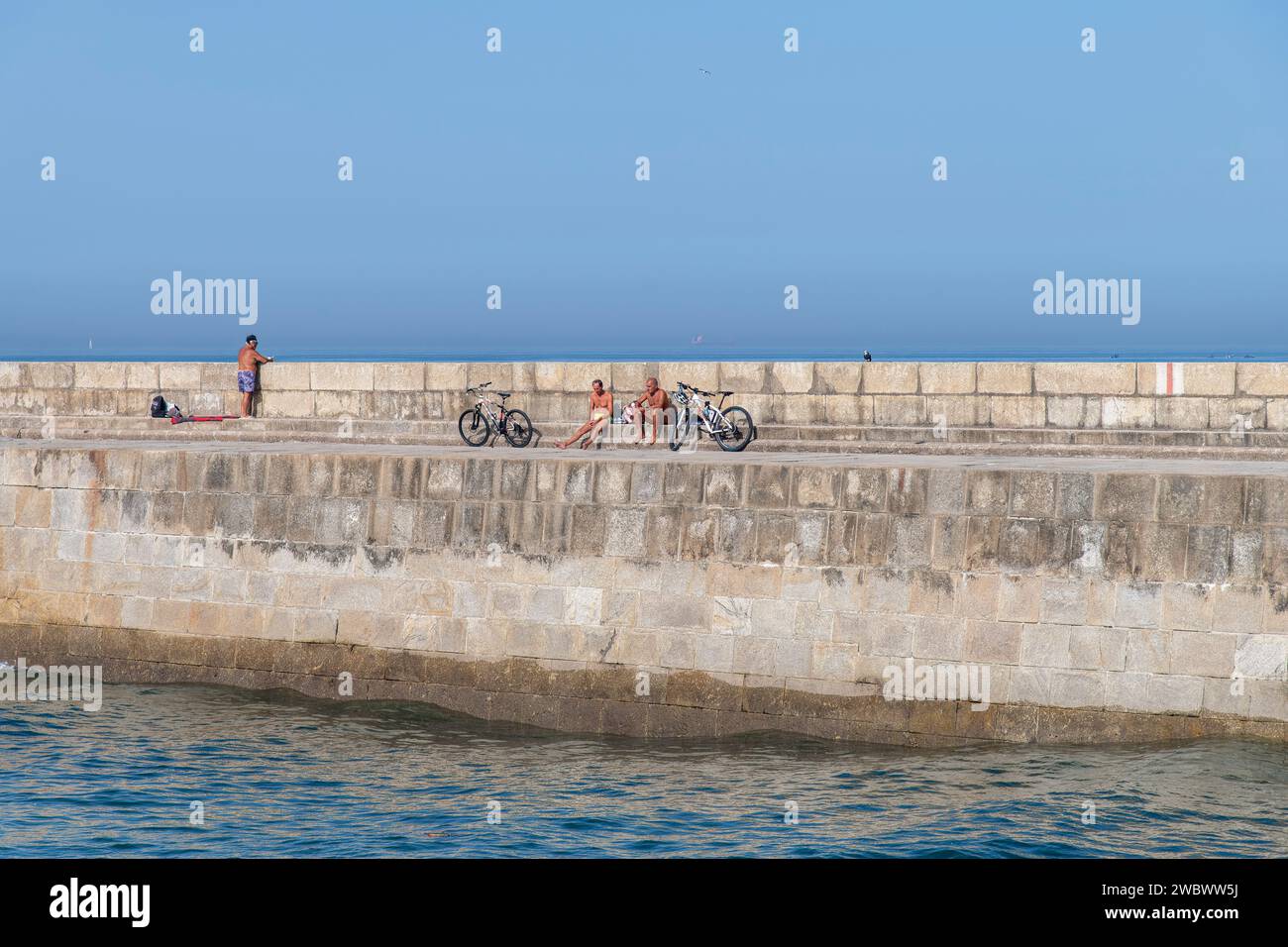 Porto, Portugal-October 5, 2022; Several man relaxing and sunbathing after bicycle ride on the concrete pier leading to Farolim da Barra do Douro Stock Photo
