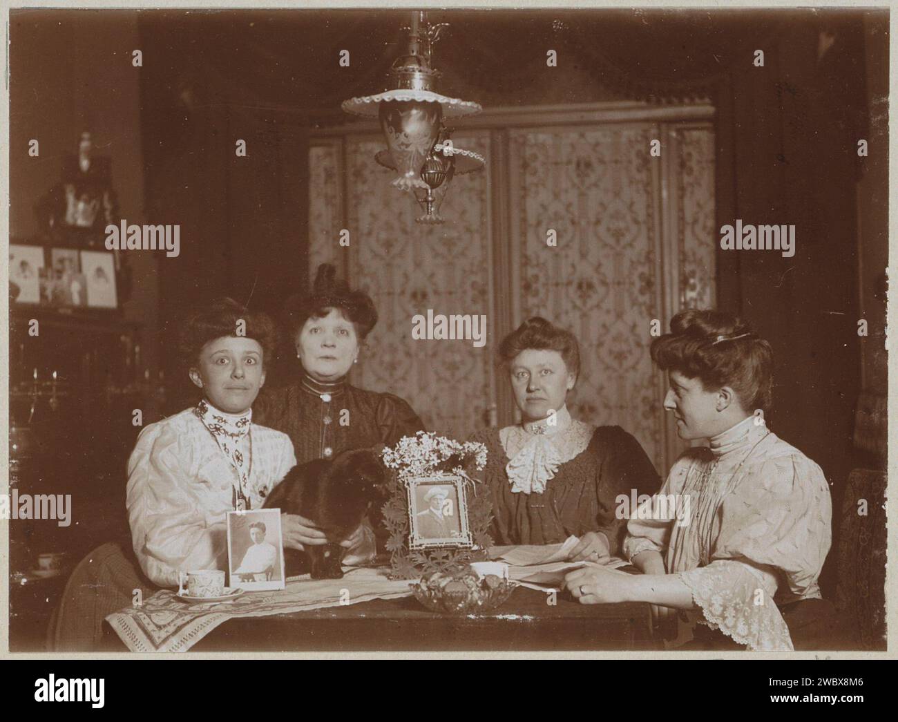 Four women at a table with two photo portraits and a cat, anonymous, c. 1900 - c. 1910 photograph Part of Family Album with photos of Wijnhandel Kraaij & Co. Bordeaux-Amsterdam.  photographic support  interior of the house. photograph. cat Stock Photo
