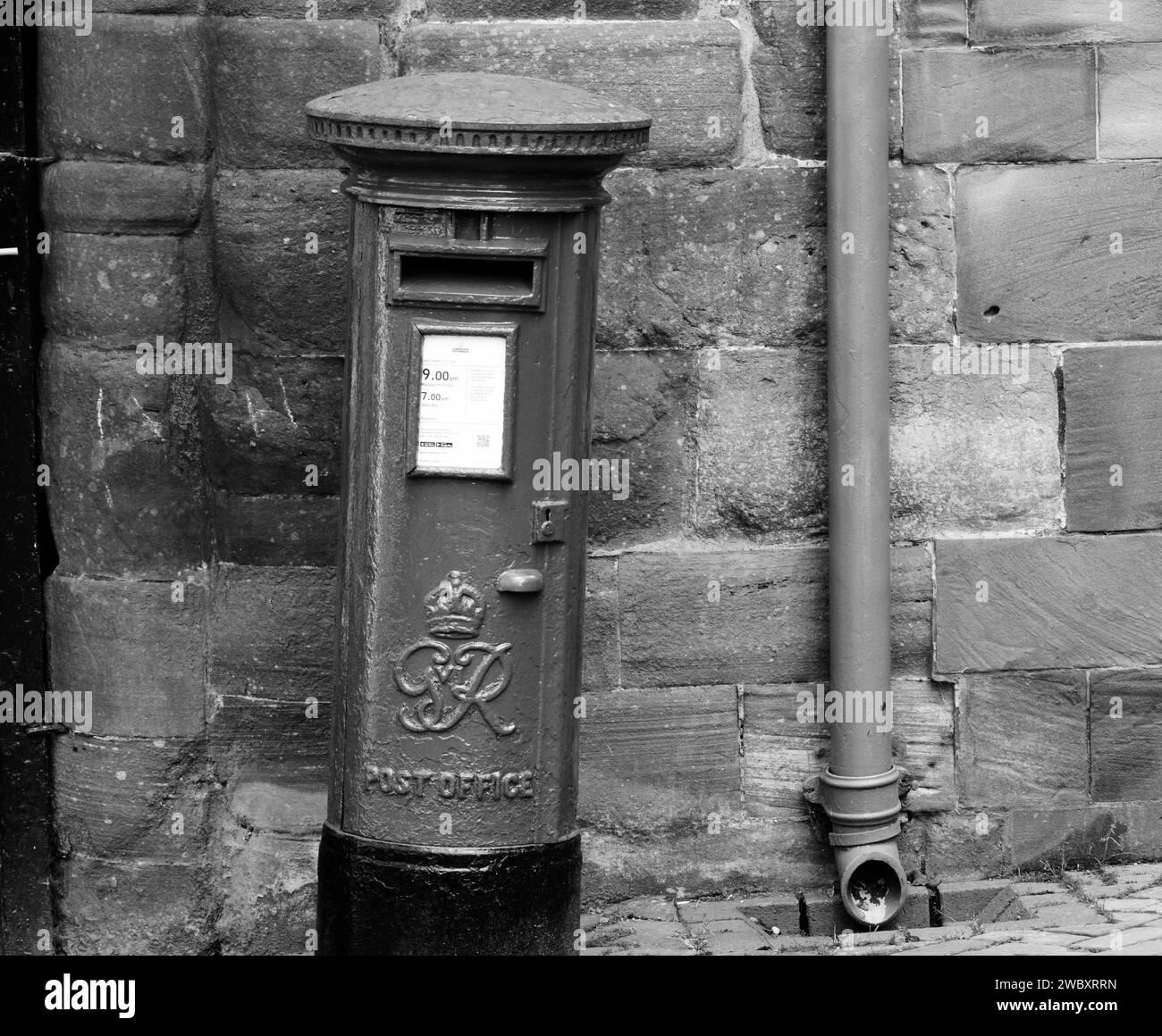 Black and white image of a post box, which is leaning to the side, against a stone wall with a drainpipe. Stock Photo
