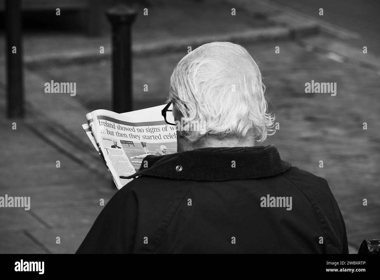 Ross On Wye, Herefordshire, England Sept 24 2023: Man with grey hair and glasses sat down reading a newspaper on a public bench. Black and white photo Stock Photo