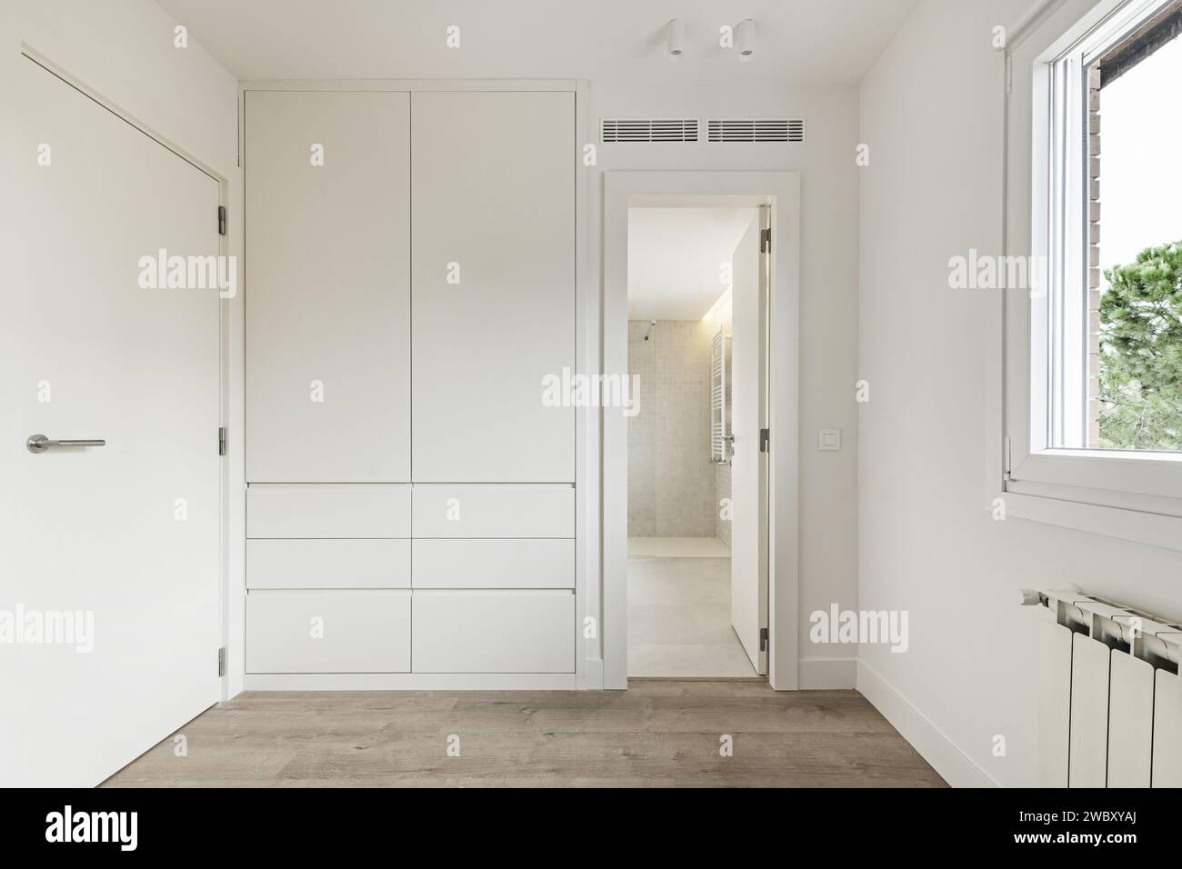 A walk-in closet next to an en-suite bathroom with white cabinet drawers and doors and white oak flooring Stock Photo