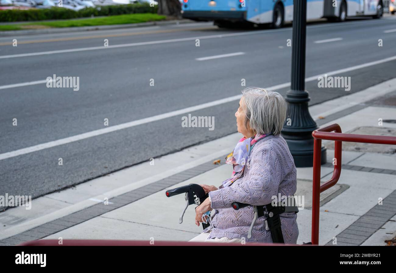 Senior woman sitting on a mobility walker outside the retirement home, taking a rest by the roadside. Stock Photo