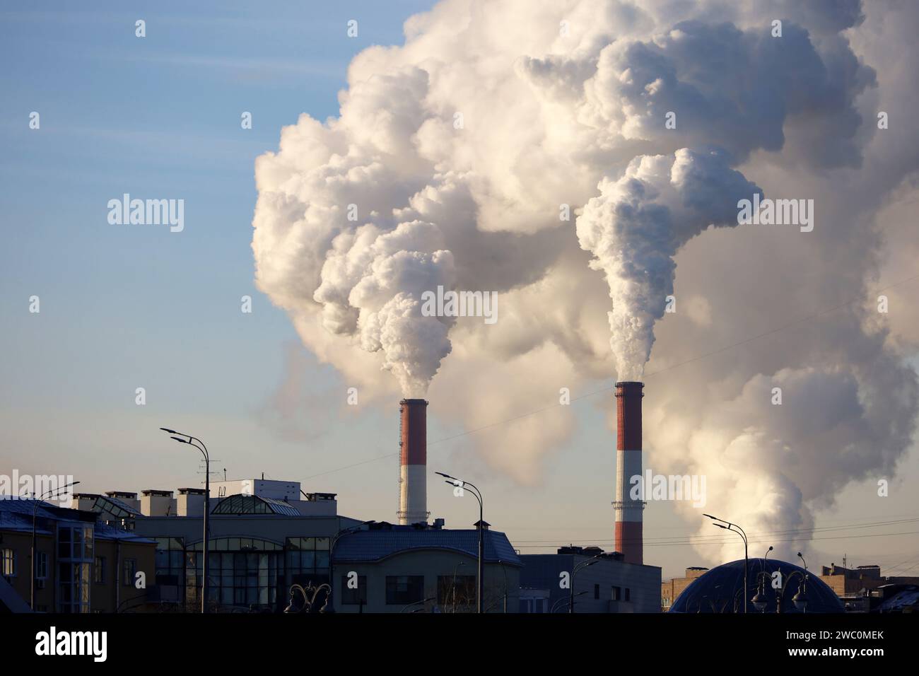 Factory chimneys with white smoke on blue sky background above the city buildings. Air pollution, heating season in winter Stock Photo