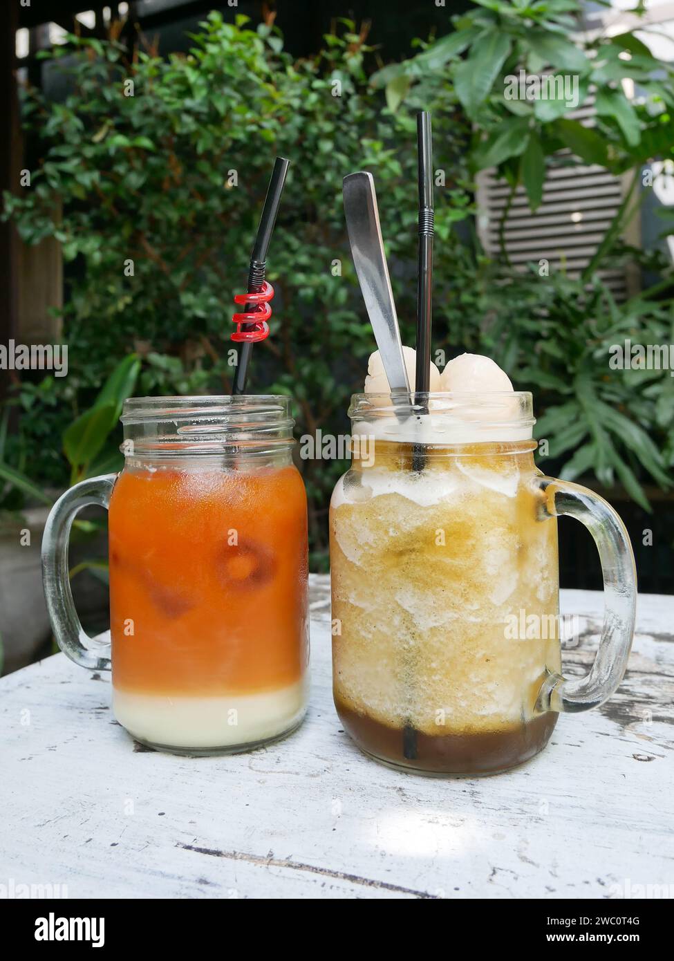 Iced lychee tea and Iced Thai tea served on a white table with a background of tropical plants and old rustic teak wood. Stock Photo