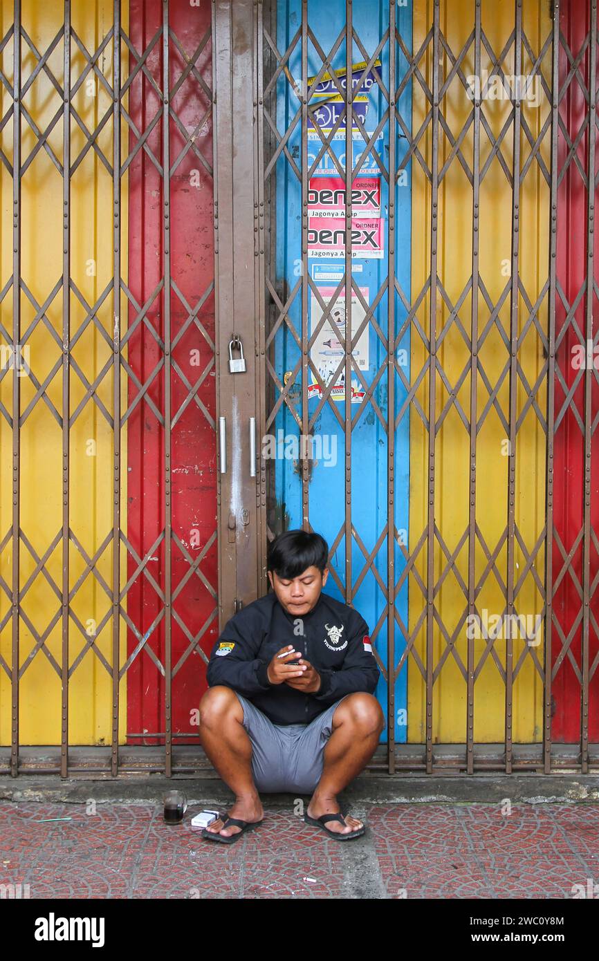 A man crouched on the ground in front of a colorful shophouse and smoking a cigarette in Indonesia. Stock Photo