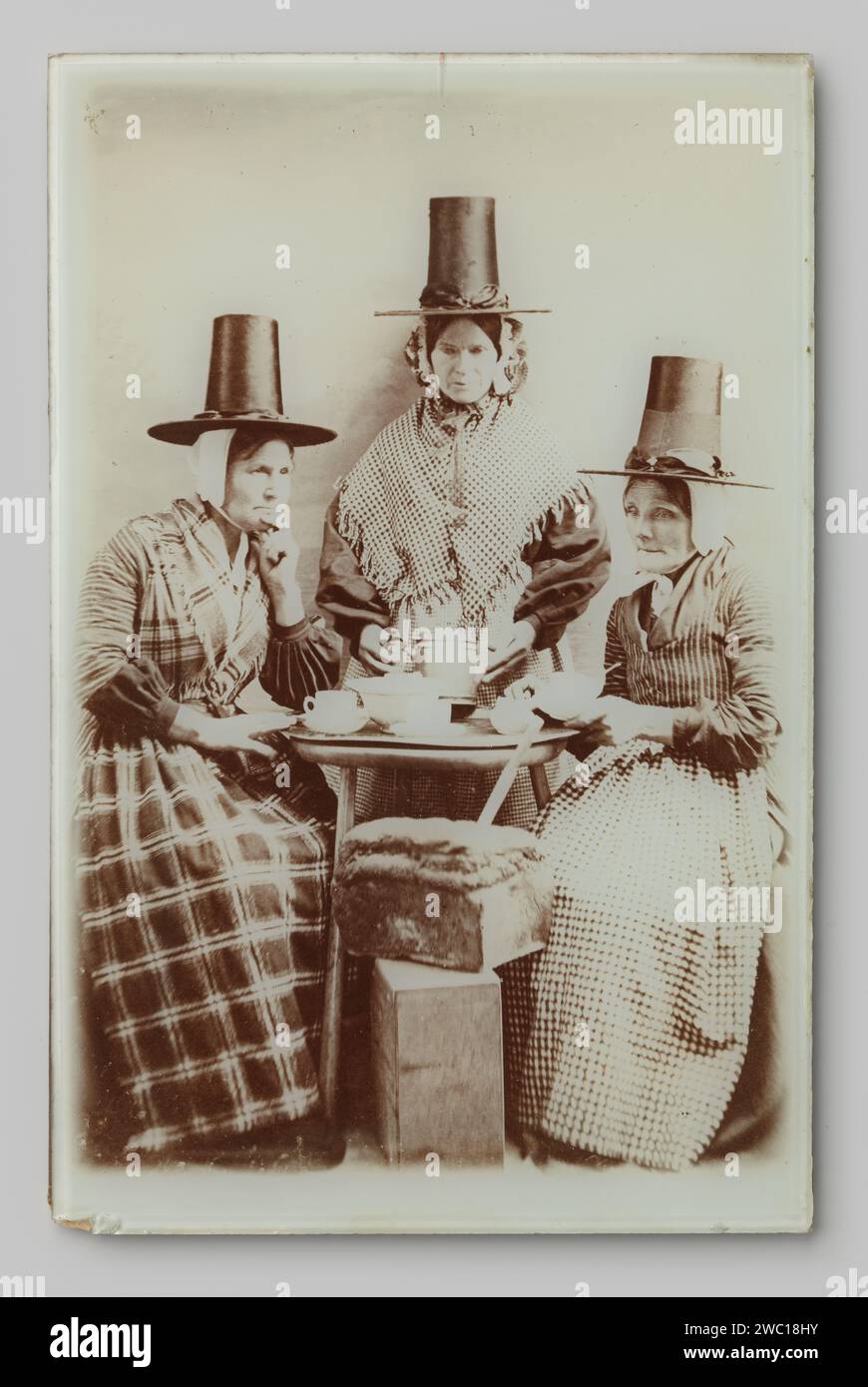 Group portrait of three unknown women at a tea table in Hindelooper Costume, Anonymous, 1890 - 1920 photograph. frame   glass. baryta paper. cardboard. metal  adult woman. anonymous historical persons portrayed in a group, in a group-portrait Stock Photo