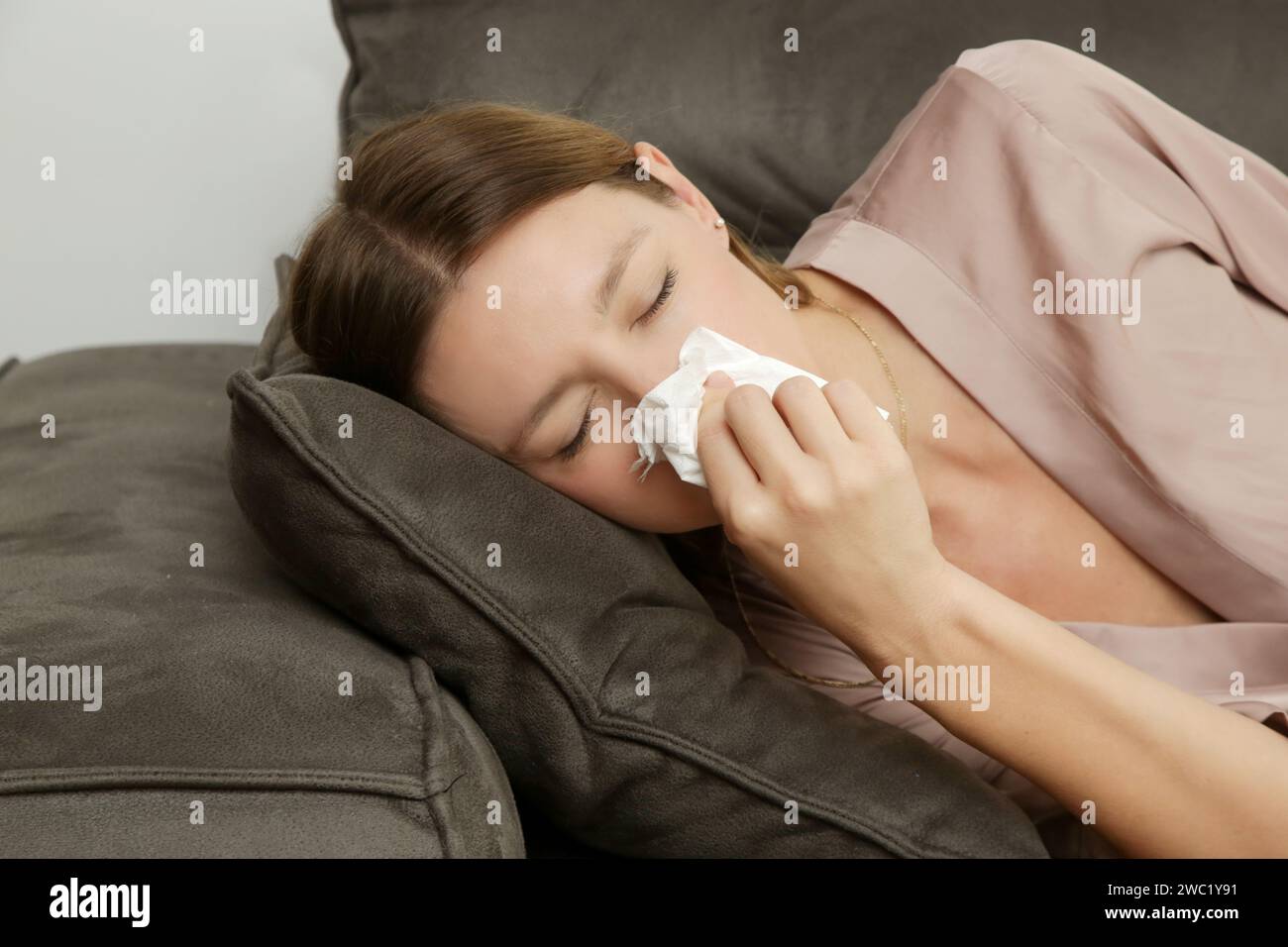 Young woman on the sofa blowing nose into a white paper tissue. Girl with allergy symptoms sneezing into a tissue. Flu, cold or allergy symptom Stock Photo