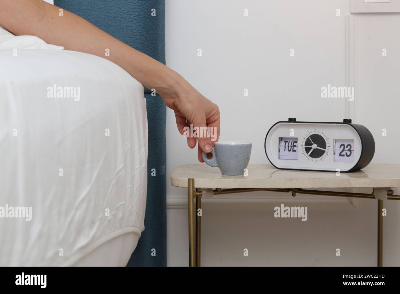 Woman in bed reaching for her first morning coffee from the nightstand. A light blue espresso cup on the bedside table next to the alarm clock Stock Photo