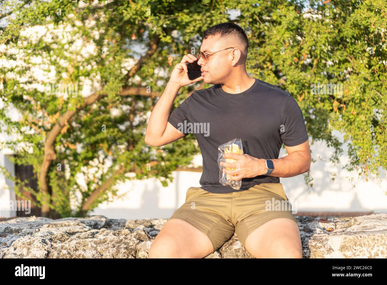 Latin Man Multitasking with Phone Call and Snack Outdoors Stock Photo