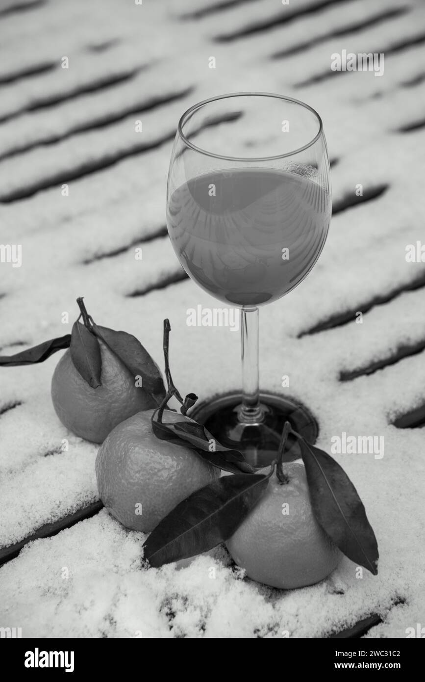 Glass of organic fresh squeezed mandarin juice on snow covered table in garden and tangerine fruits. Healthy eating lifestyle background. Black white. Stock Photo