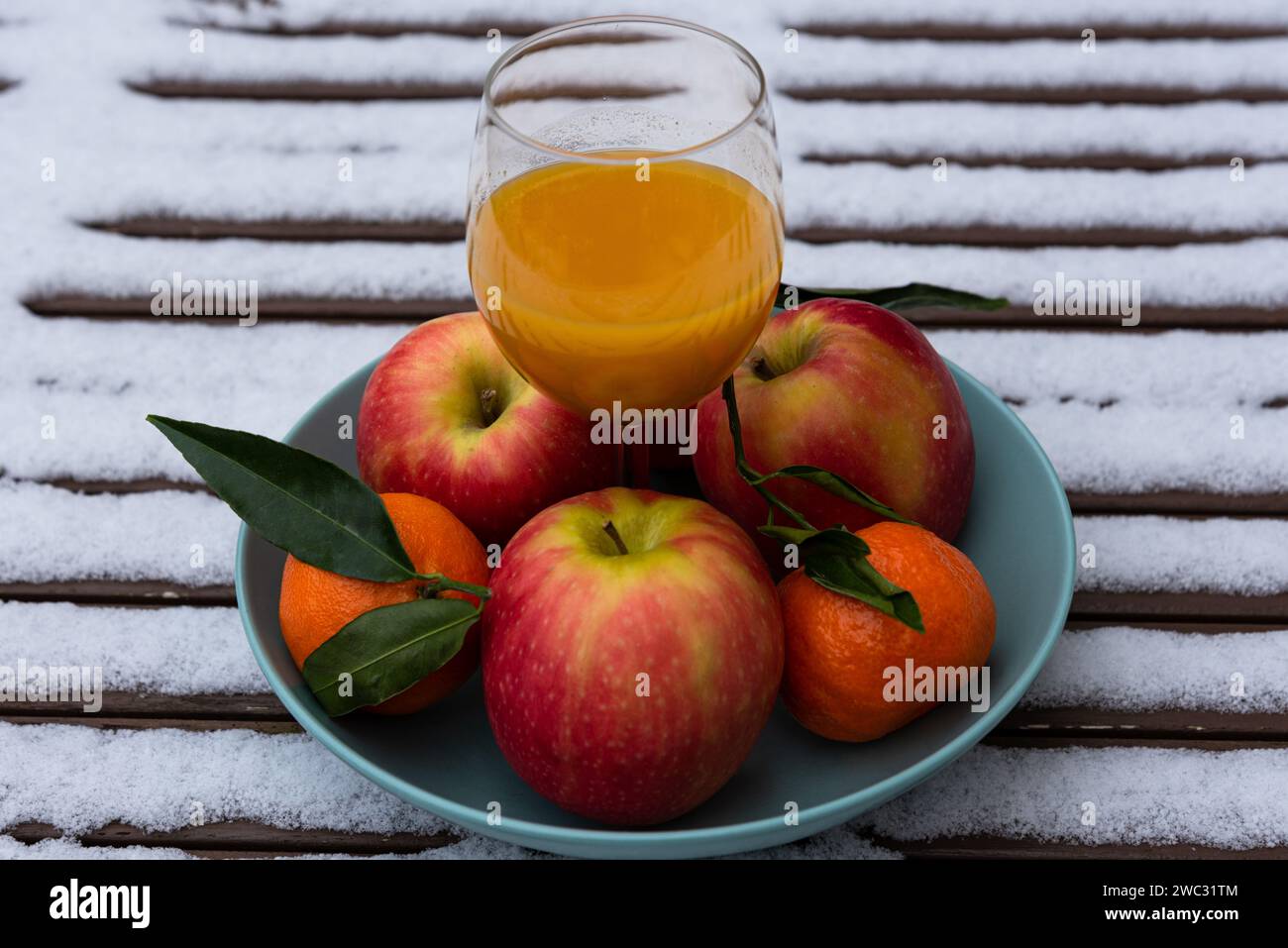 Glass of organic fresh squeezed fruit juice on snow covered table in garden and tangerines with apples in bowl. Healthy eating lifestyle background. Stock Photo