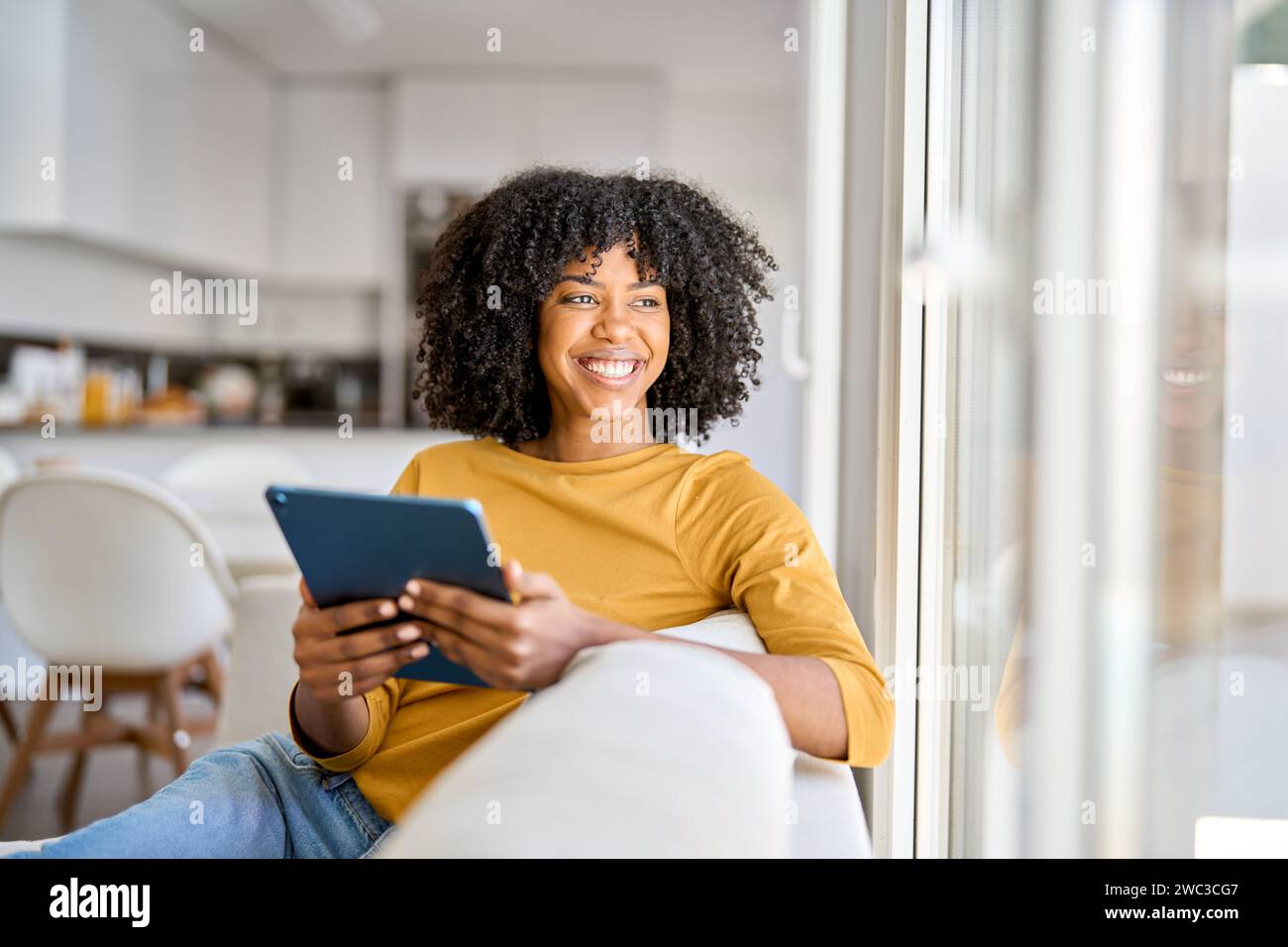 Happy African American lady using tab device looking away on sofa at home. Stock Photo