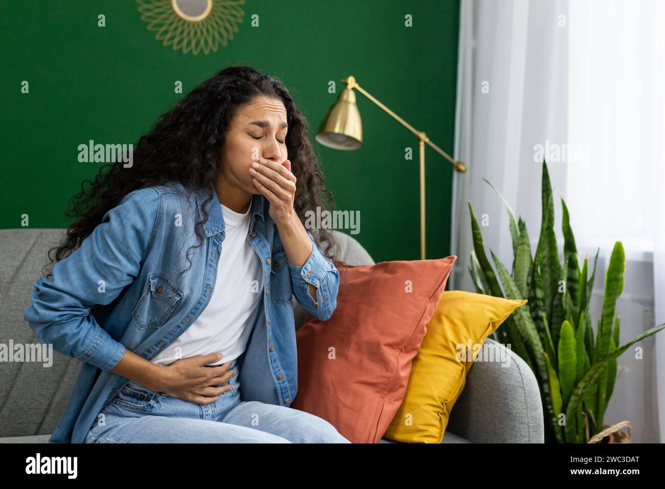 A distressed young woman feeling nauseous on a couch, hand over her mouth, trying to suppress the urge to vomit. Stock Photo