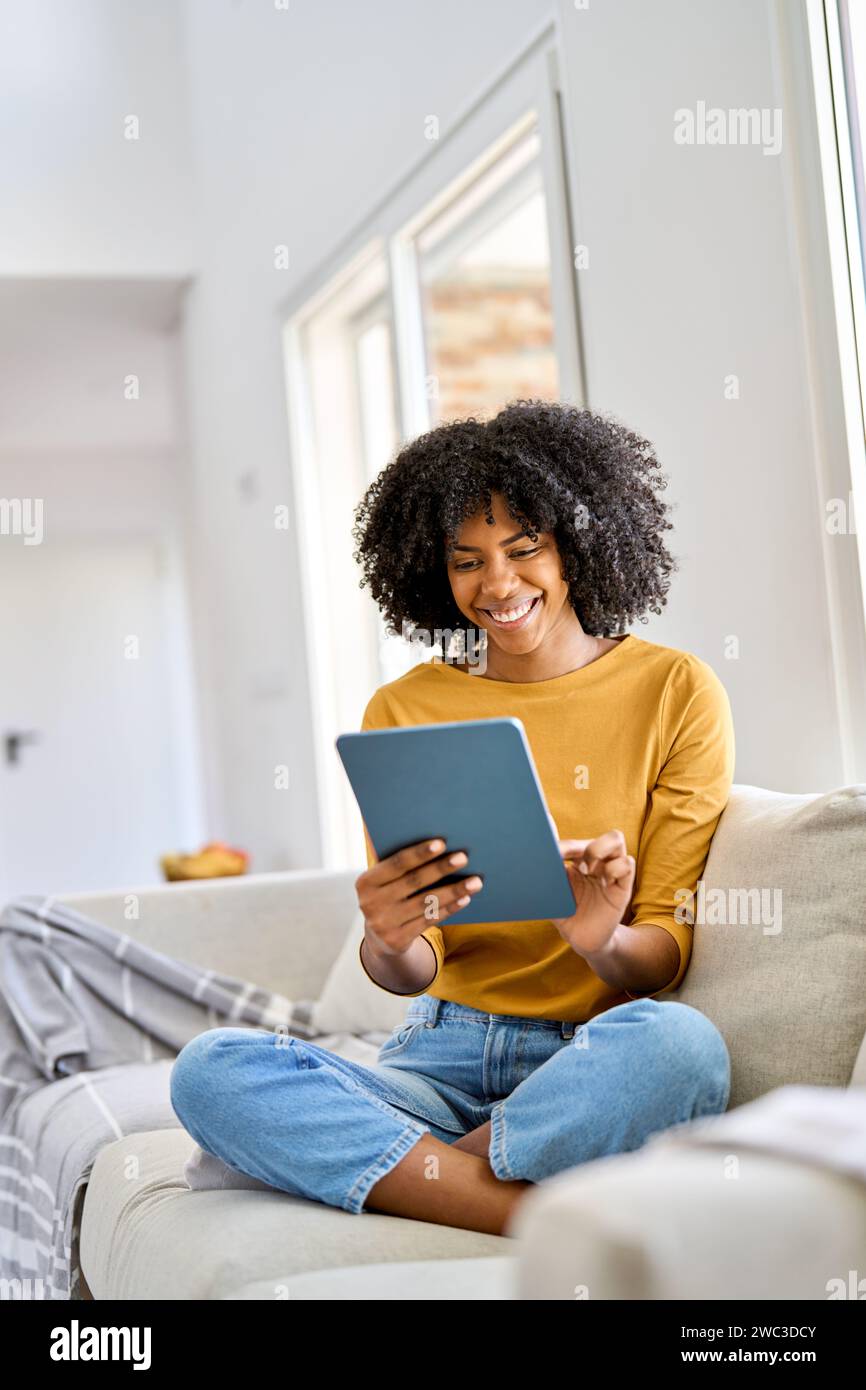 Smiling African American girl sitting on sofa at home using digital tablet. Stock Photo