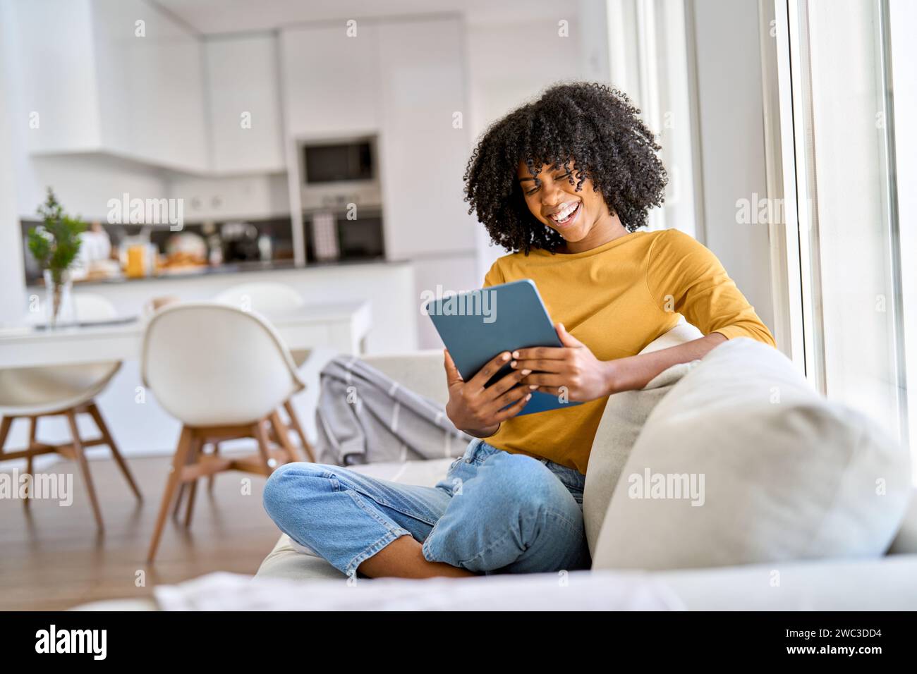 Happy young African woman using digital tablet laughing on sofa at home. Stock Photo