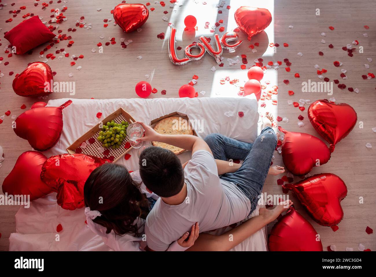 Top view of young couple sharing casual, intimate moment together on bed with red heart shaped balloons. Man and woman embracing each others in Valent Stock Photo