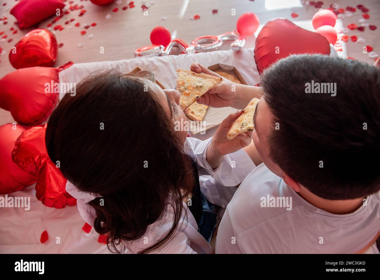 Top view of young couple feeding each other, enjoying pizza from takeaway box, with glasses of champagne, green grapes. Man and woman sharing festive, Stock Photo