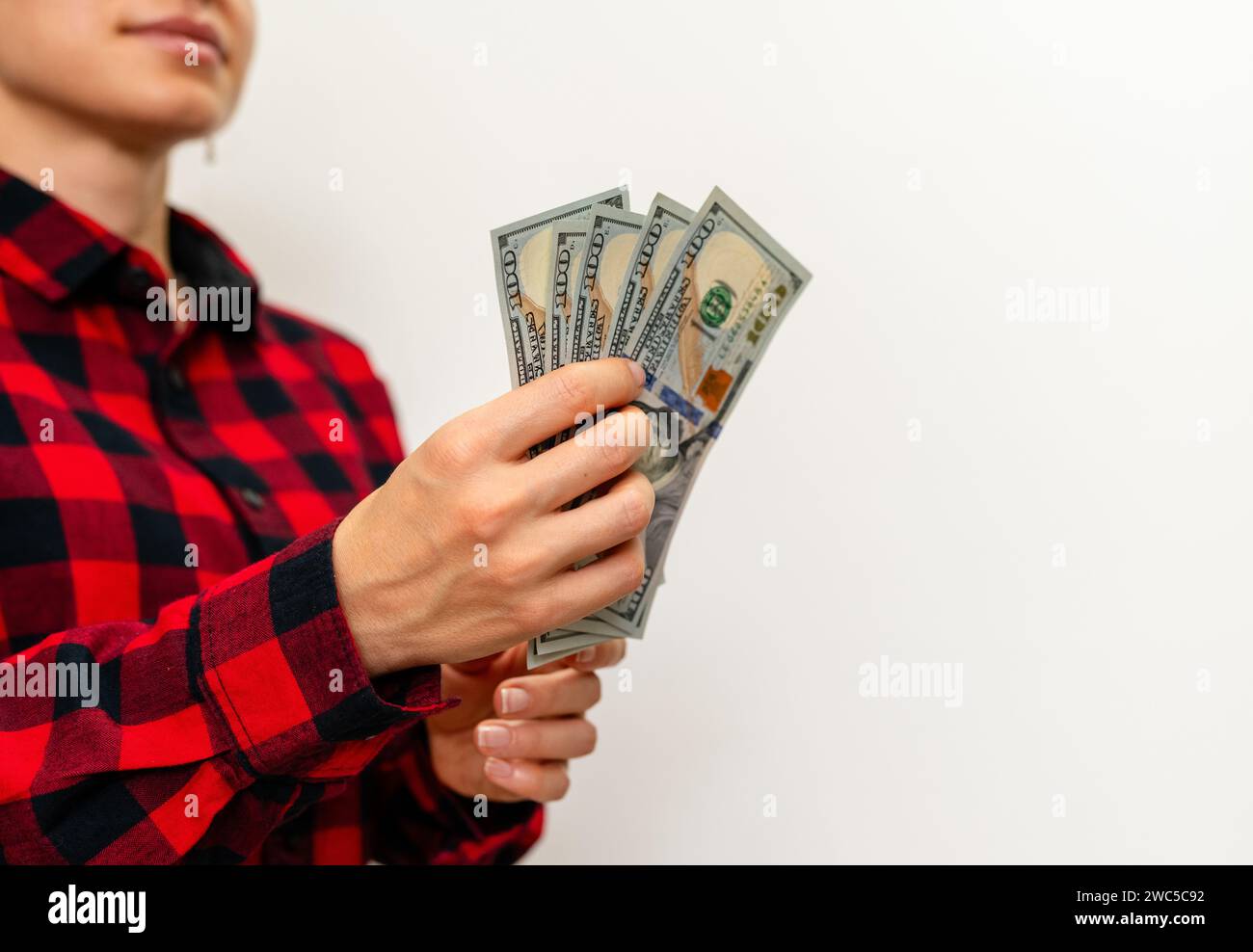 Female person in red casual shirt holds 100 dollar bills in hand in front of plain background with copy space. Stock Photo
