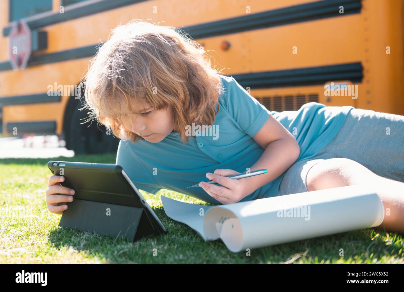 Child does school homework laying on grass in the park near school bus ...