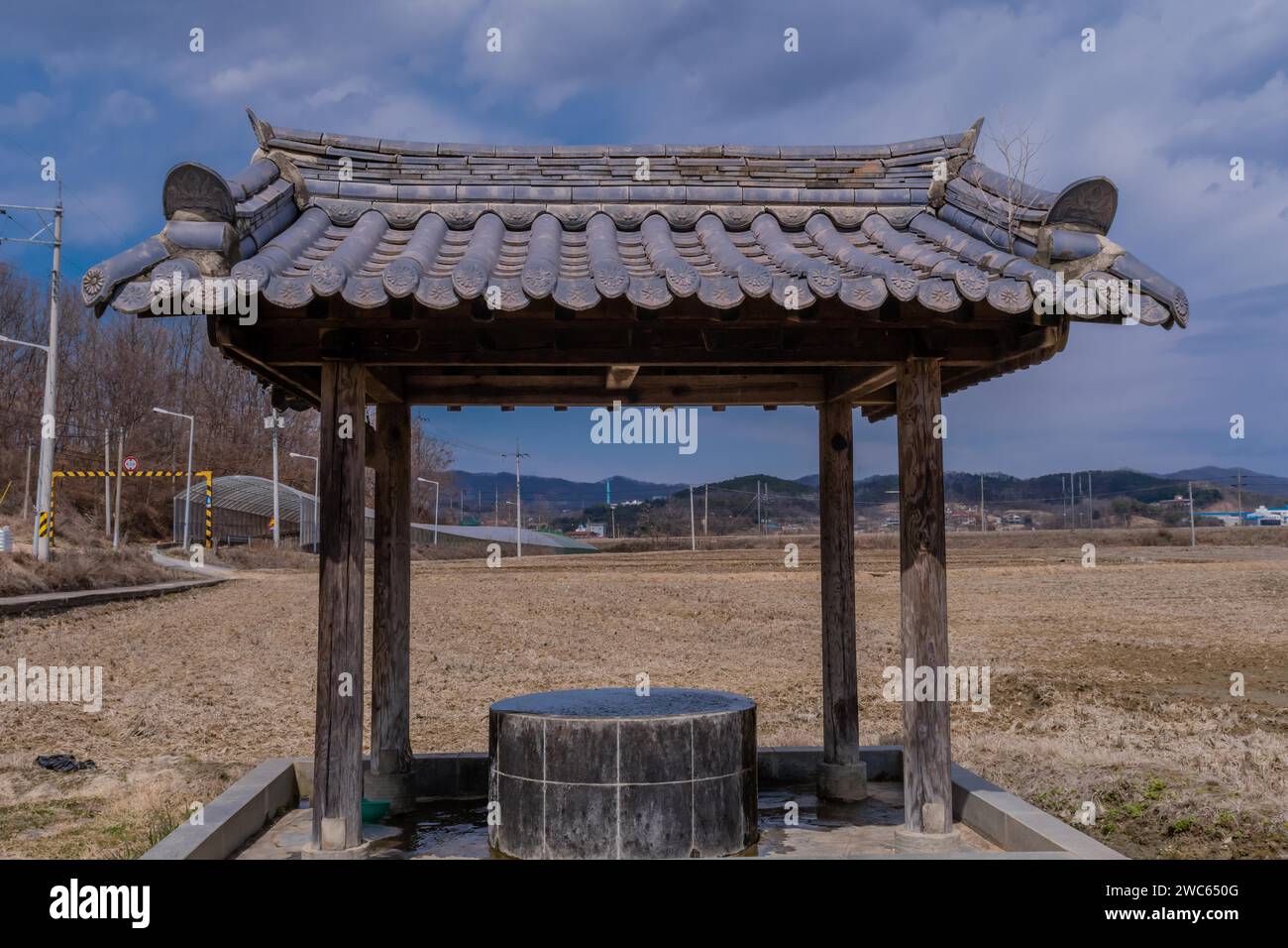 Covered water well under ceramic tiled roof full of water in a mountainside public park with plowed field in background Stock Photo