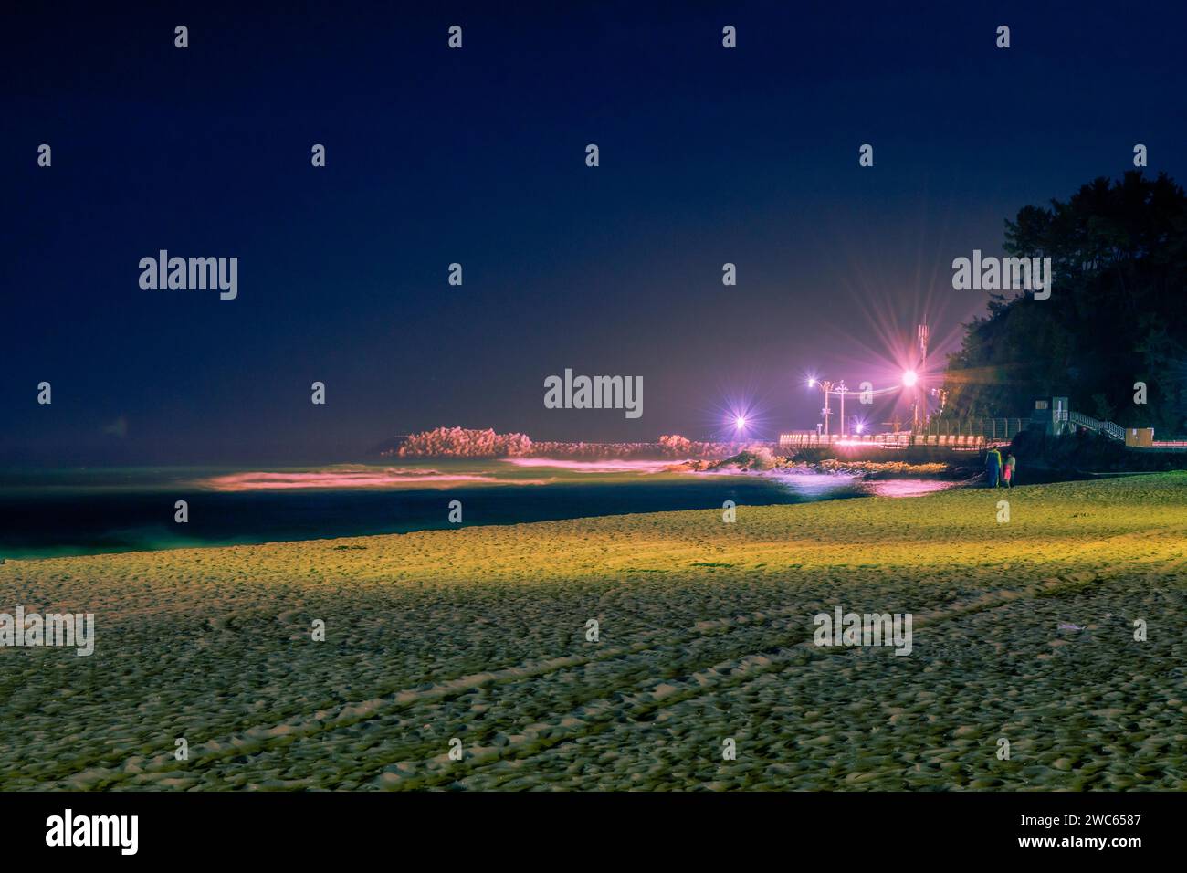 Night scene at beach bathed in soft light with unrecognizable people and pier lit up by street lights in background Stock Photo