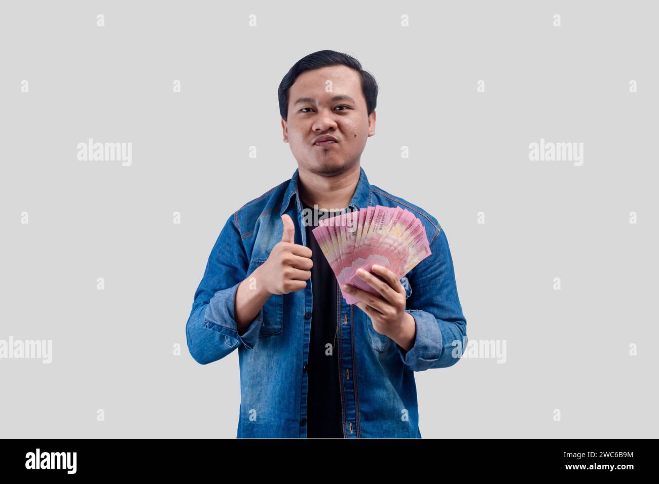 Young Asian man showing arrogant face expression while holding paper money and give a thumbs up Stock Photo