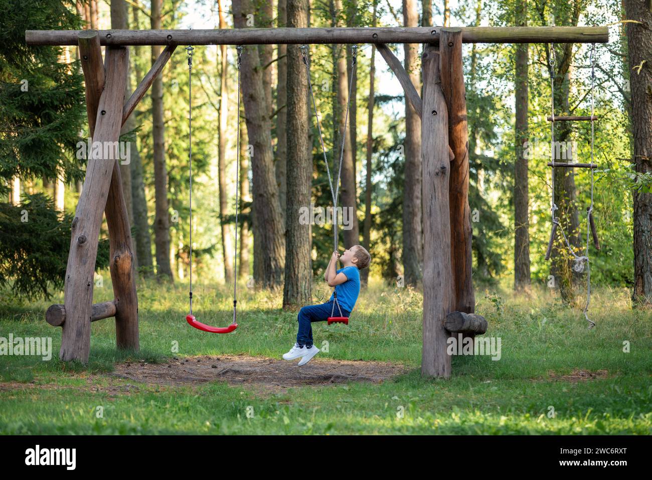 A little boy sits on a large rope swing in a sunny forest. The child plays with the ropes, twisting them and then quickly spins around its axis. Stock Photo