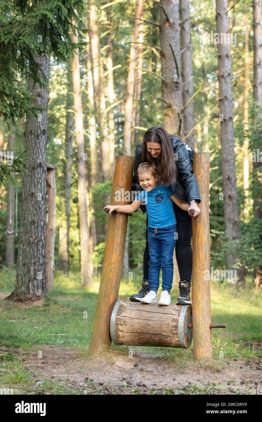 A mother and her little son have fun on a rotating wooden barrel - forest attraction. The wooden footpath equipment in the forest is designed for runn Stock Photo