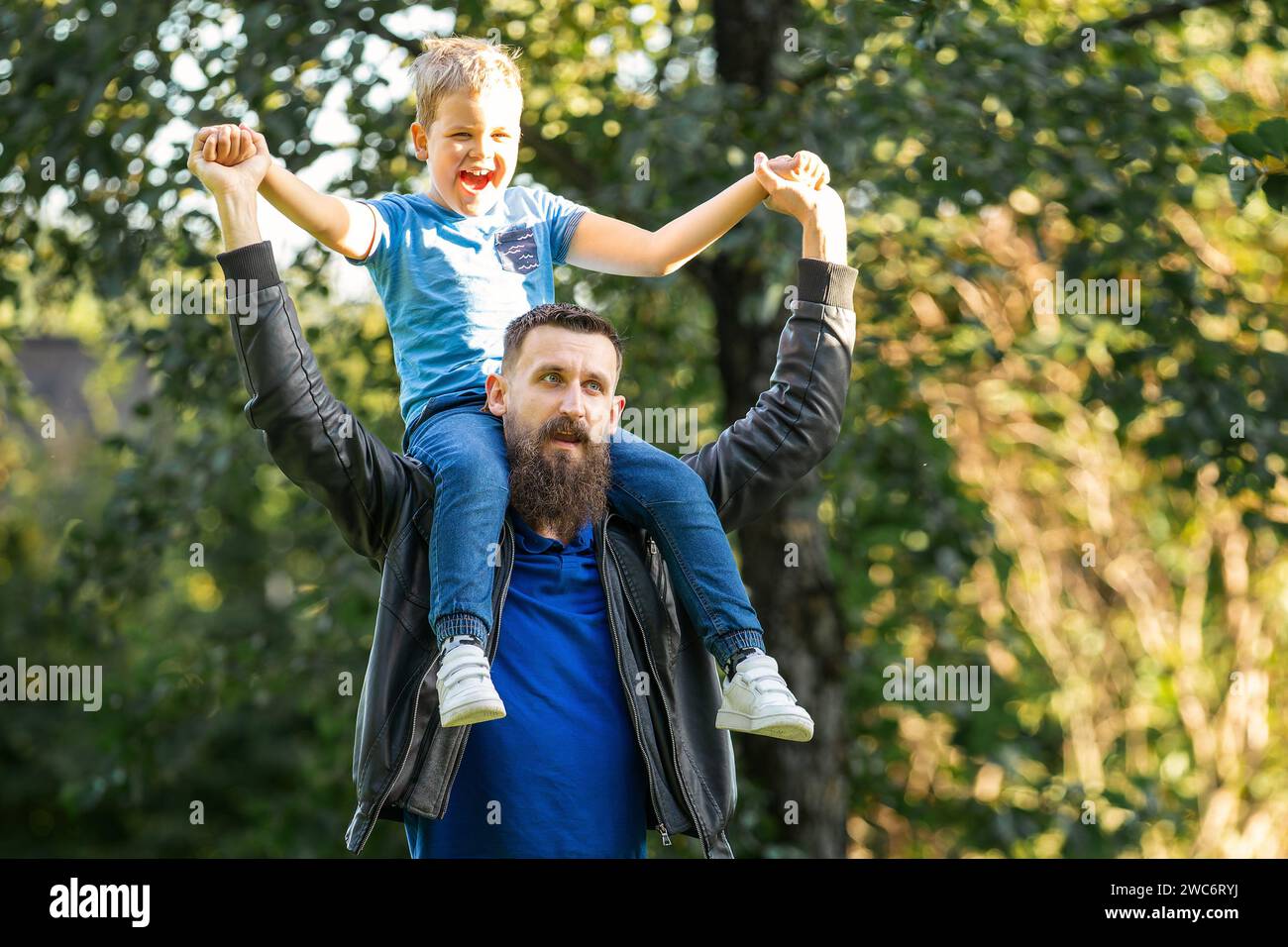 Portrait of a happy laughing sunlit child on the shoulders of his bearded father against a background of summer foliage. Stock Photo