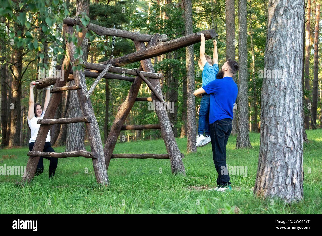 A mother, father and her little son are having fun in the summer in the forest on a large wooden swing. The mother jokes and lifts her son high, tippi Stock Photo
