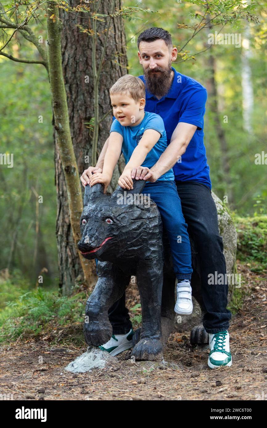 Vertical portrait of father and son riding on a black wooden wolf sculpture in the forest. Stock Photo