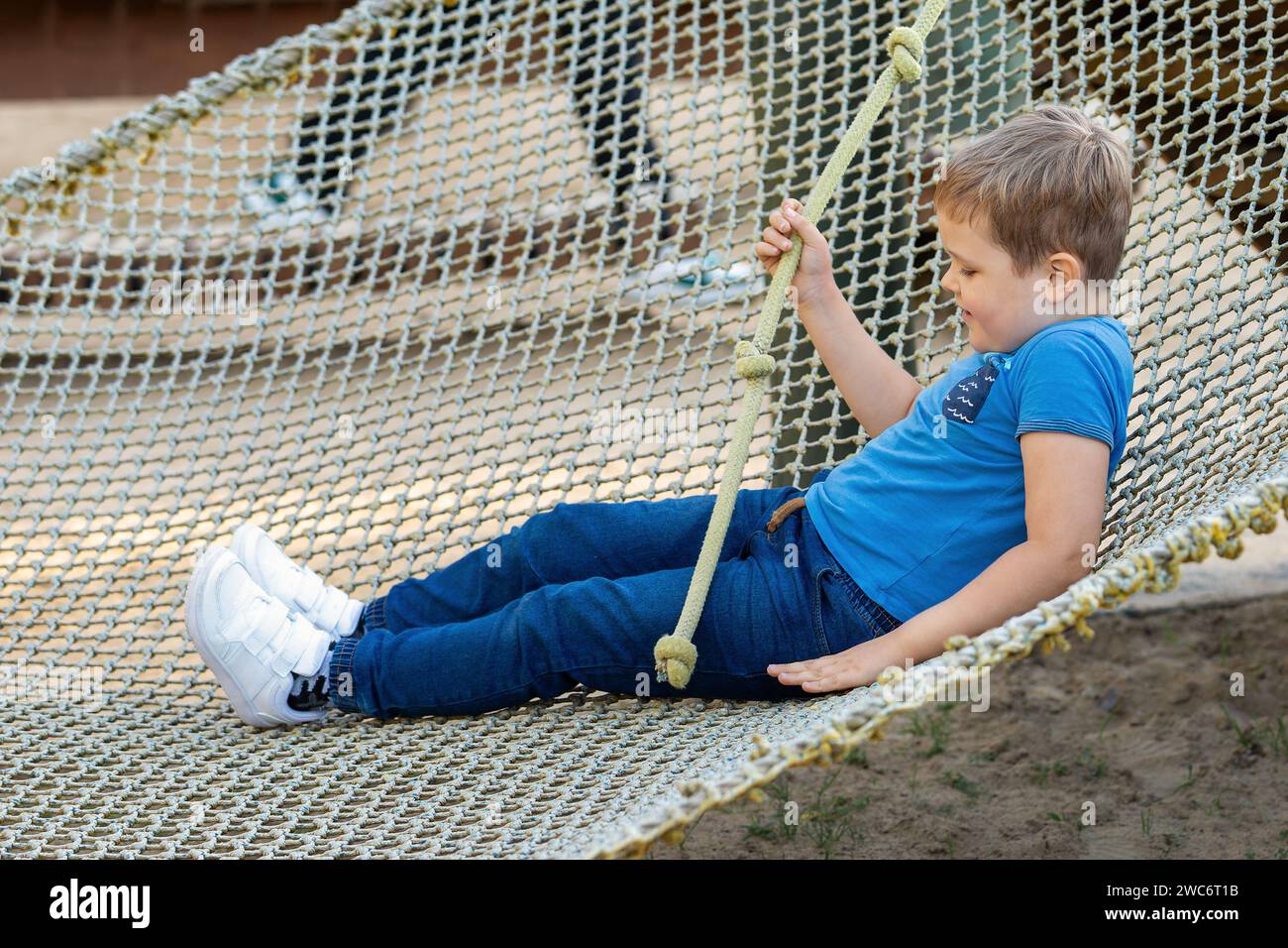 Portrait of a little boy in climbing gear in a rope park, holding a rope with a knot. Kid plays in rope adventure park. Obstacle course for children. Stock Photo