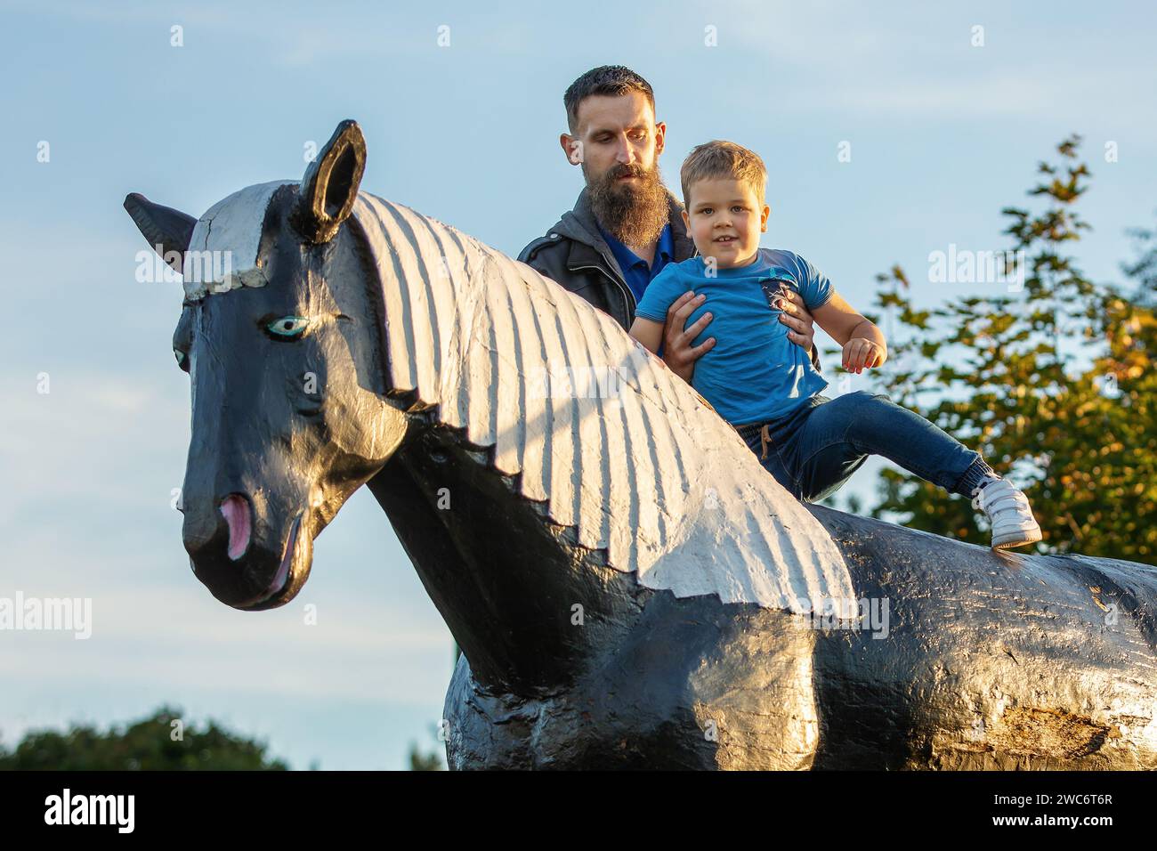 Father put up his son he helps him climb onto the large horse figure in the outdoor park Stock Photo