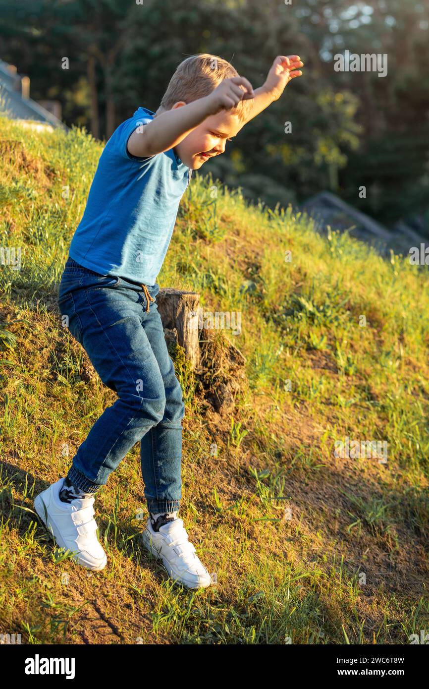 Backlit by the evening sun, a happy child runs down a steep grassy slope with arms raised high. Stock Photo
